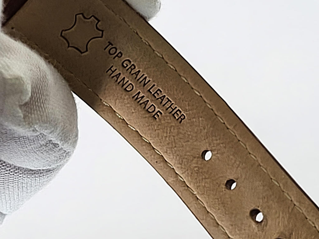A close-up of a tan leather watch strap for a Reliance wristwatch, held by a white gloved hand, showing the interior side with 'TOP GRAIN LEATHER HAND MADE' text and a leather stamp icon.