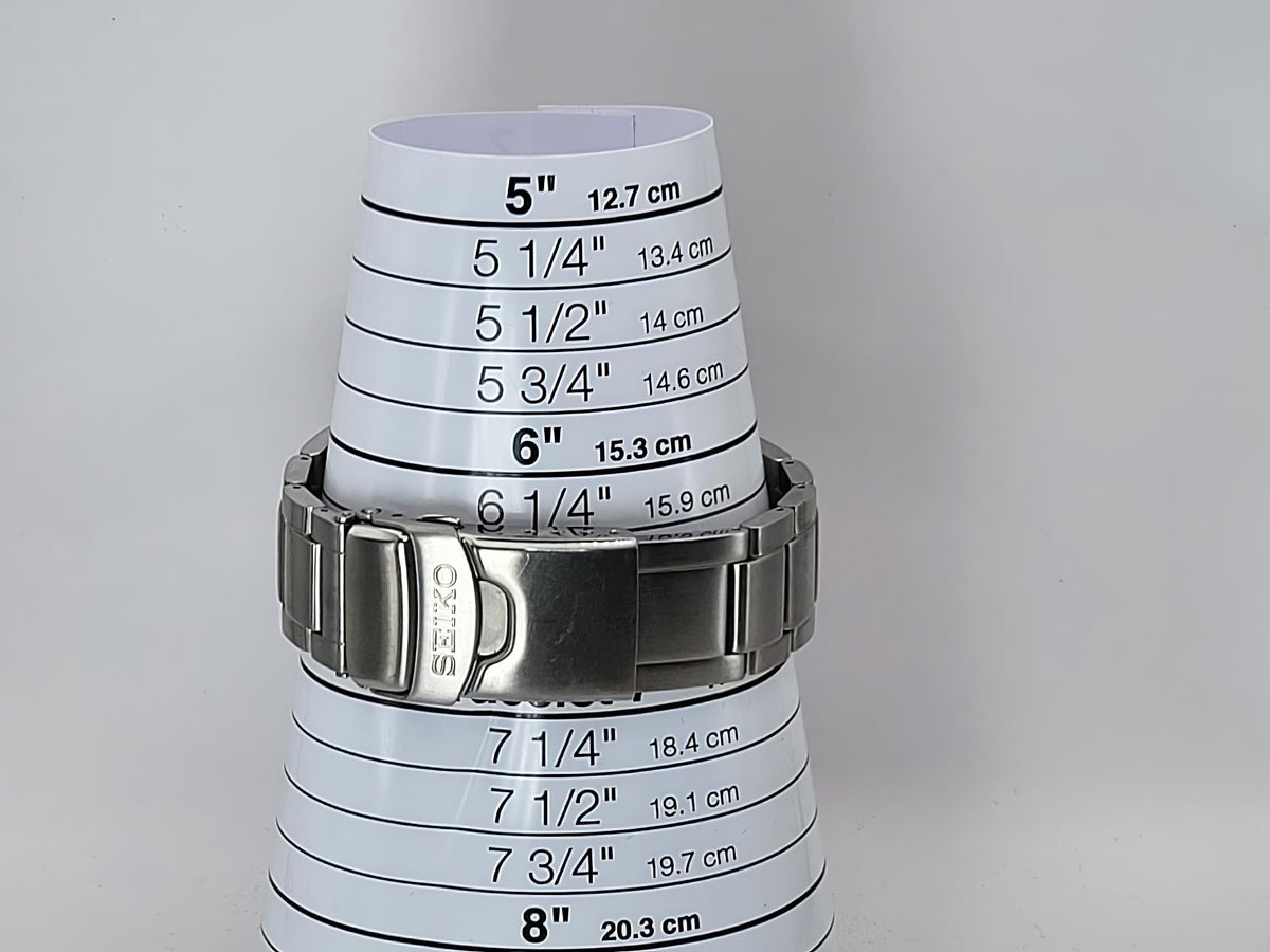 A stainless steel Seiko Flightmaster watch band wrapped around a white plastic cone measuring chart printed with size conversions in inches and centimeters.
