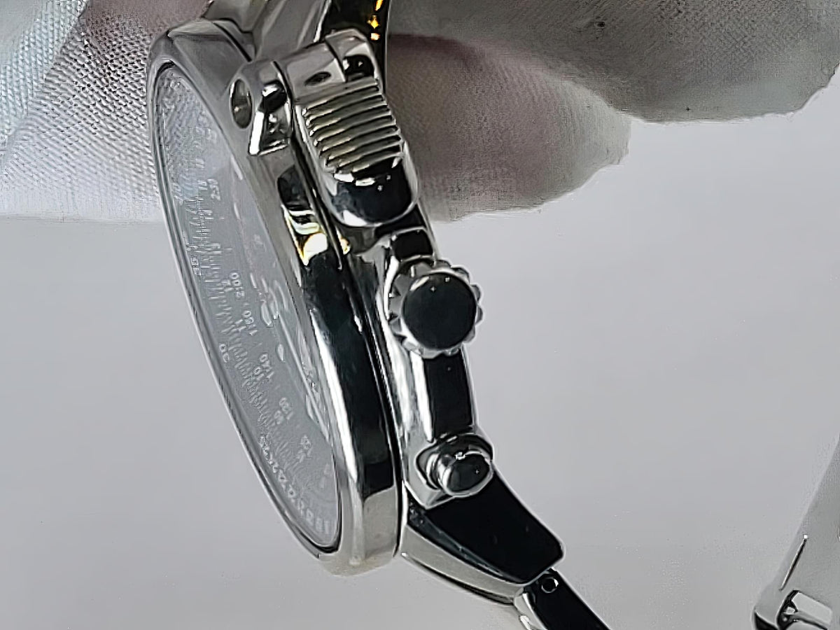 Close-up side view of a stainless steel Seiko Flightmaster wristwatch with a silver case, black crown and pushers, and a white dial, held by a white-gloved hand against a light gray background.