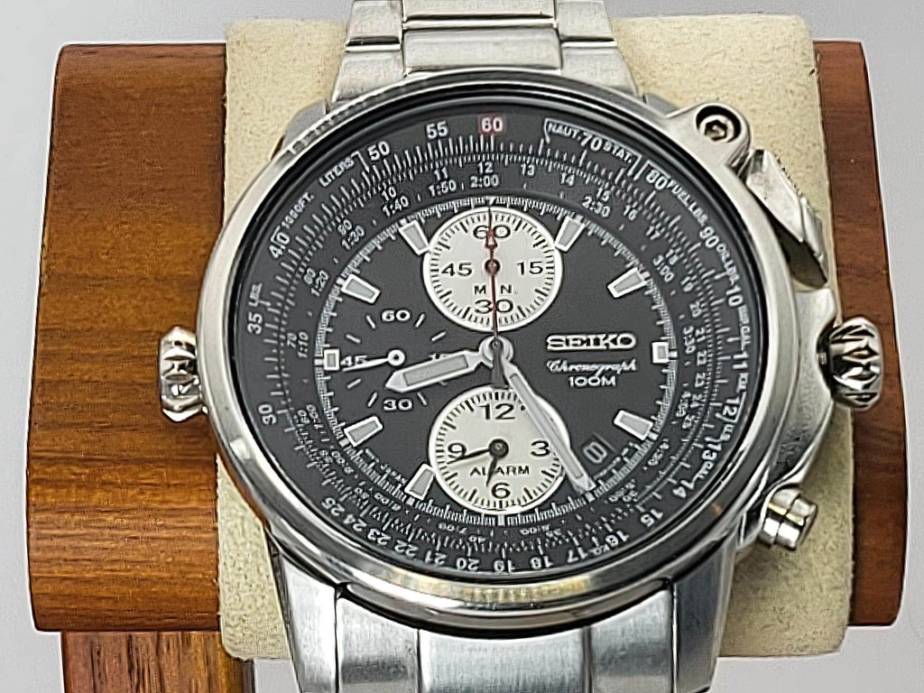 A Seiko Flightmaster watch with a silver stainless steel case and link bracelet, featuring a black dial with white sub-dials and a tachymeter bezel, displayed on a wooden stand with a beige cushion.