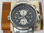A Seiko Flightmaster watch with a silver stainless steel case and link bracelet, featuring a black dial with white sub-dials and a tachymeter bezel, displayed on a wooden stand with a beige cushion.