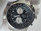 A close-up of a Seiko Flightmaster watch with a black dial, silver stainless steel case, and white-gloved hands, resting on a white textured surface.