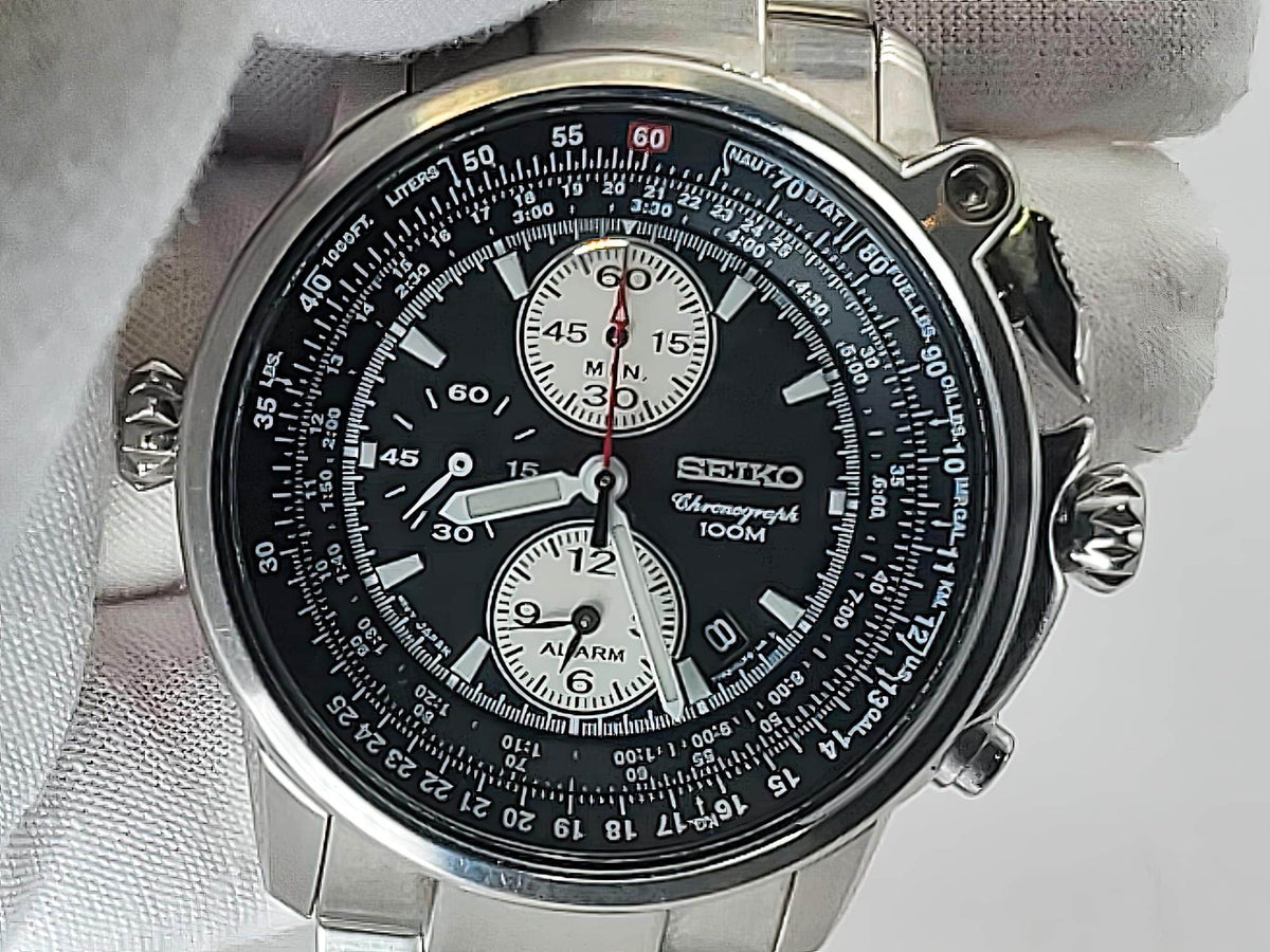 A close-up of a Seiko Flightmaster watch with a black dial, silver stainless steel case, and white-gloved hands, resting on a white textured surface.