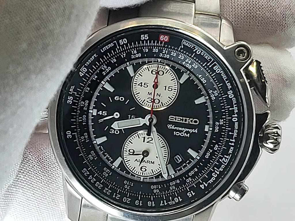 A close-up of a Seiko Flightmaster wristwatch with a black dial, silver stainless steel case, and white subdials, resting on a white textured surface.