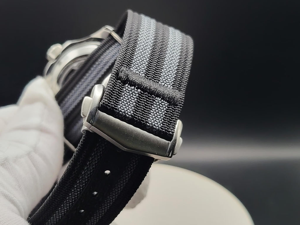 Close-up of The Alias SJT Original wristwatch strap with a woven black and gray pattern, attached to a silver metal buckle, held by a white gloved hand against a dark background.