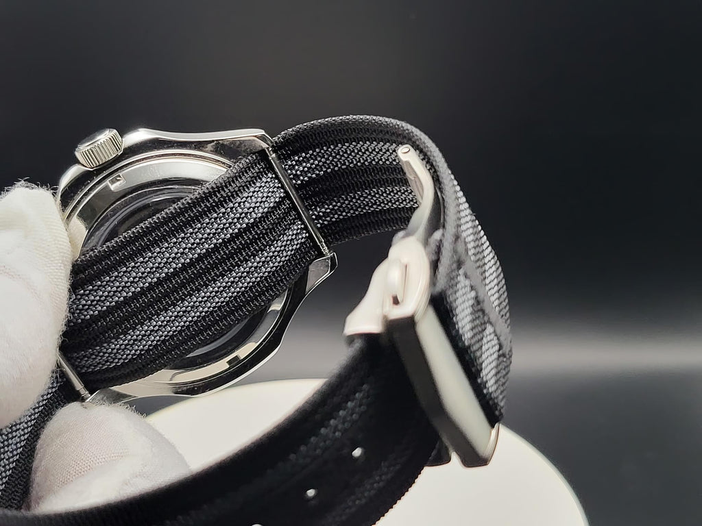 Close-up of The Alias SJT Original wristwatch strap with a black and gray striped woven fabric and a silver buckle, held by a white gloved hand against a dark background.