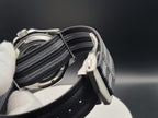 Close-up of The Alias SJT Original wristwatch strap with a black and gray striped woven fabric and a silver buckle, held by a white gloved hand against a dark background.