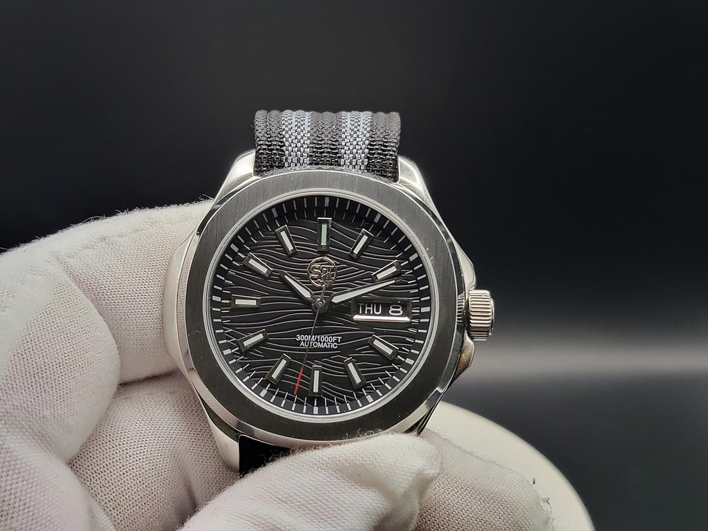 A close-up of The Alias SJT Original wristwatch with a dark textured dial, silver-toned case, and gray-and-black striped fabric strap, held by a white gloved hand against a dark background.