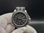A close-up of The Alias SJT Original wristwatch with a dark textured dial, silver-toned case, and gray-and-black striped fabric strap, held by a white gloved hand against a dark background.