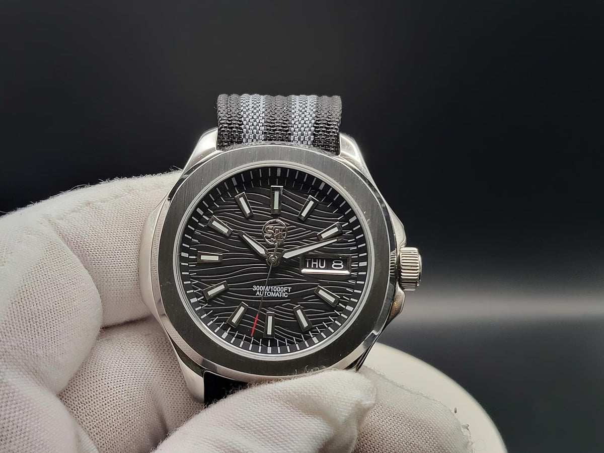 A close-up of The Alias SJT Original wristwatch with a dark textured dial, silver-toned case, and gray-and-black striped fabric strap, held by a white gloved hand against a dark background.