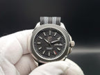 A close-up of The Alias SJT Original wristwatch with a silver-toned metal case, textured dark gray dial, and gray fabric strap, held by a white gloved hand against a dark background.