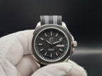 A close-up of The Alias SJT Original wristwatch with a silver-toned metal case, black dial, and textured gray-and-black fabric strap, held in a white gloved hand against a dark background.