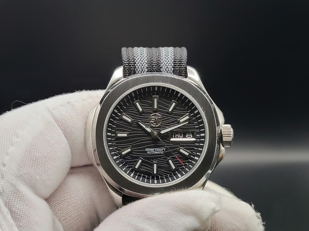 A close-up of The Alias SJT Original wristwatch with a silver-toned metal case, black dial, and textured gray-and-black fabric strap, held in a white gloved hand against a dark background.