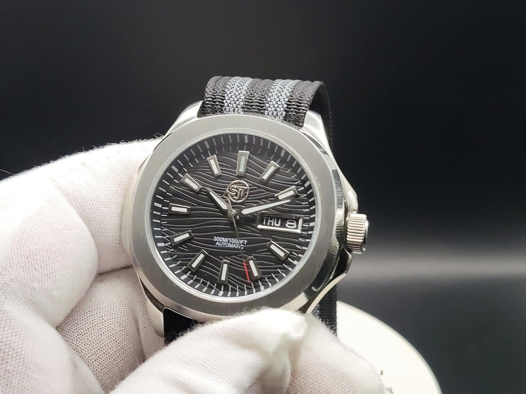 A close-up of The Alias SJT Original wristwatch with a silver-toned metal case, a dark textured dial, and a black-and-gray striped fabric strap, held by a white gloved hand against a dark background.
