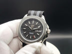 A close-up of The Alias SJT Original wristwatch with a silver-toned metal case, a dark textured dial, and a black-and-gray striped fabric strap, held by a white gloved hand against a dark background.