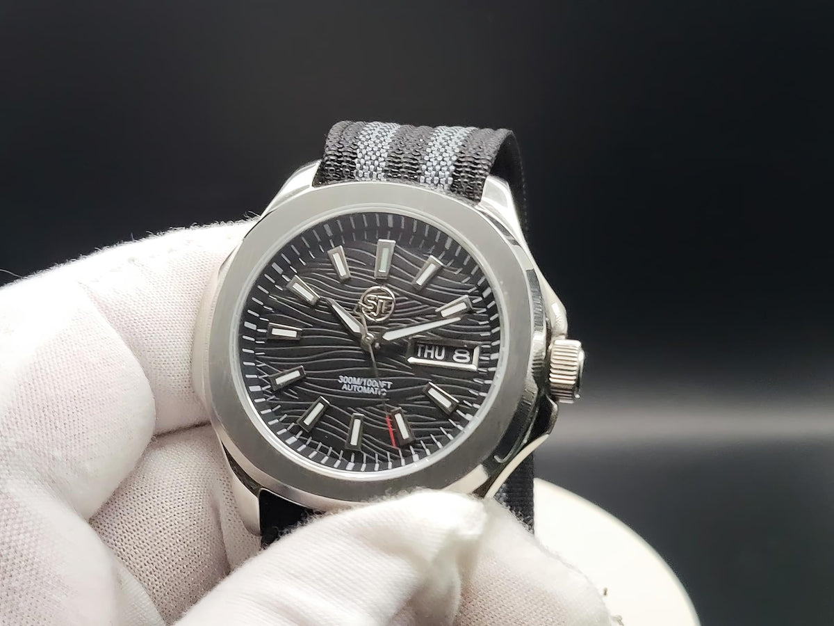 A close-up of The Alias SJT Original wristwatch with a silver-toned metal case, a dark textured dial, and a black-and-gray striped fabric strap, held by a white gloved hand against a dark background.