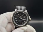 A close-up of The Alias SJT Original wristwatch with a black dial, silver-toned case, and a gray-and-black textured fabric strap, held in a white-gloved hand against a dark background.