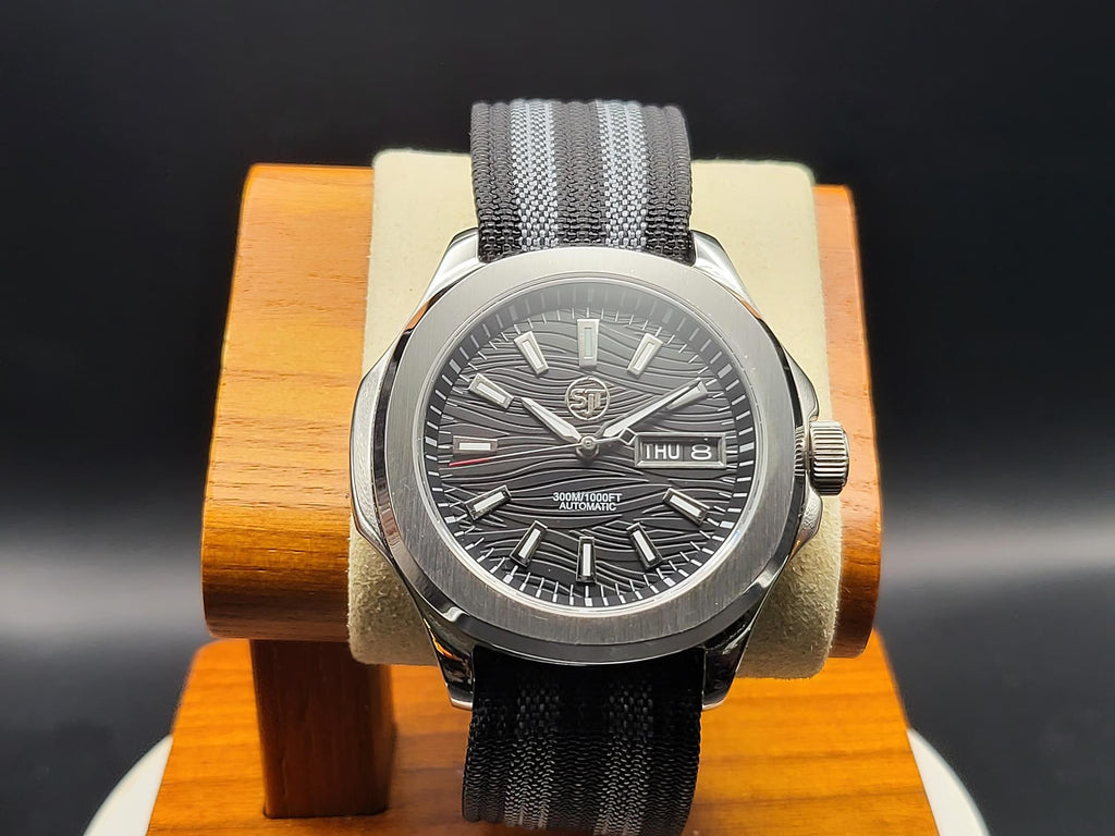The Alias SJT Original wristwatch with a dark gray wavy dial, silver-toned case, and a gray fabric strap, displayed on a wooden stand against a dark background.