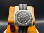 The Alias SJT Original wristwatch with a dark gray wavy dial, silver-toned case, and a gray fabric strap, displayed on a wooden stand against a dark background.