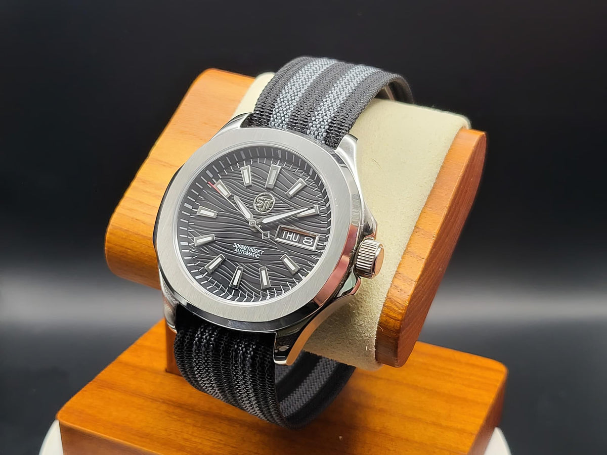 The Alias SJT Original wristwatch with a silver-toned metal case, a textured dark gray dial, and a gray fabric strap, displayed on a wooden stand against a dark background.
