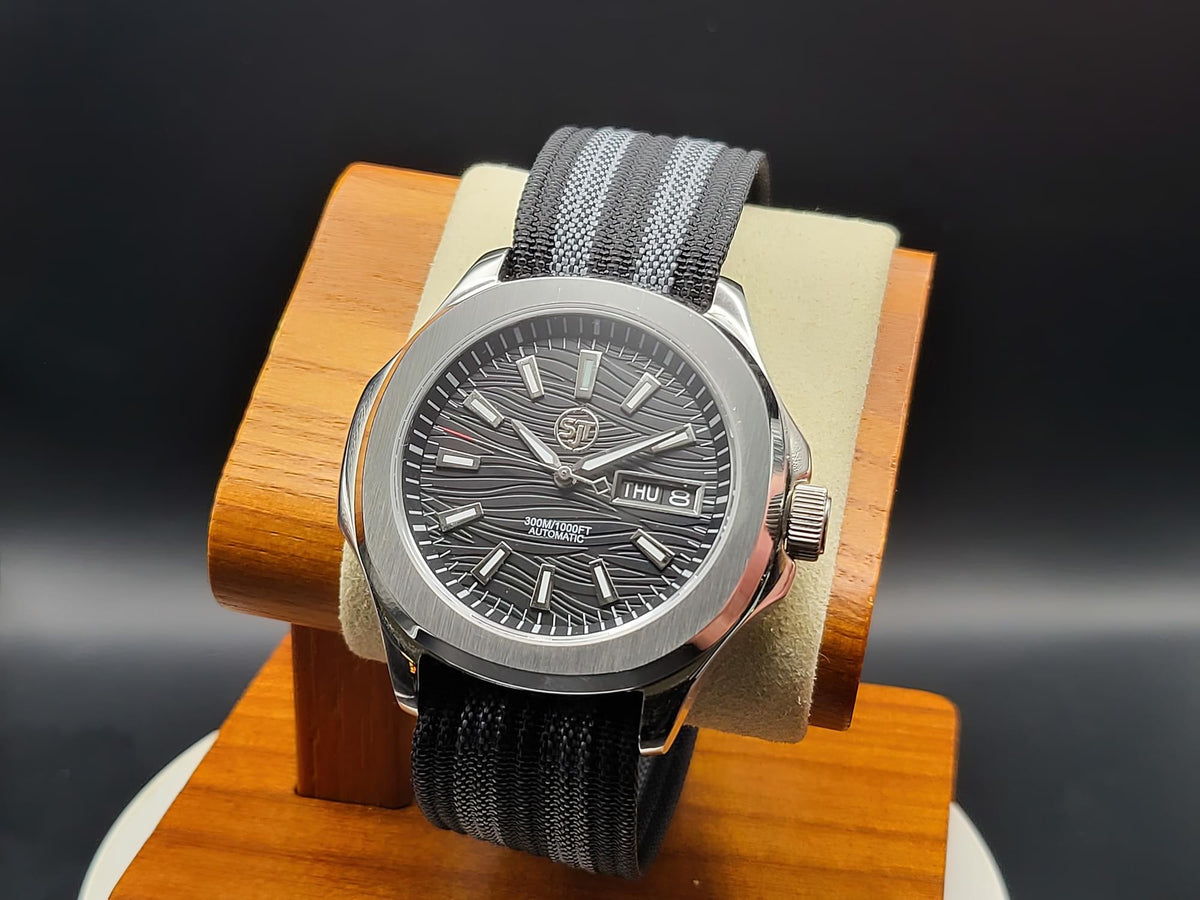 The Alias SJT Original wristwatch with a dark gray textured dial, silver-toned case, and a gray-and-black striped fabric strap, displayed on a wooden stand against a dark background.
