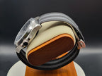 The Alias SJT Original wristwatch, a silver-toned stainless steel watch with a gray and black woven fabric strap, displayed on a beige cushion with a wooden base against a dark background.