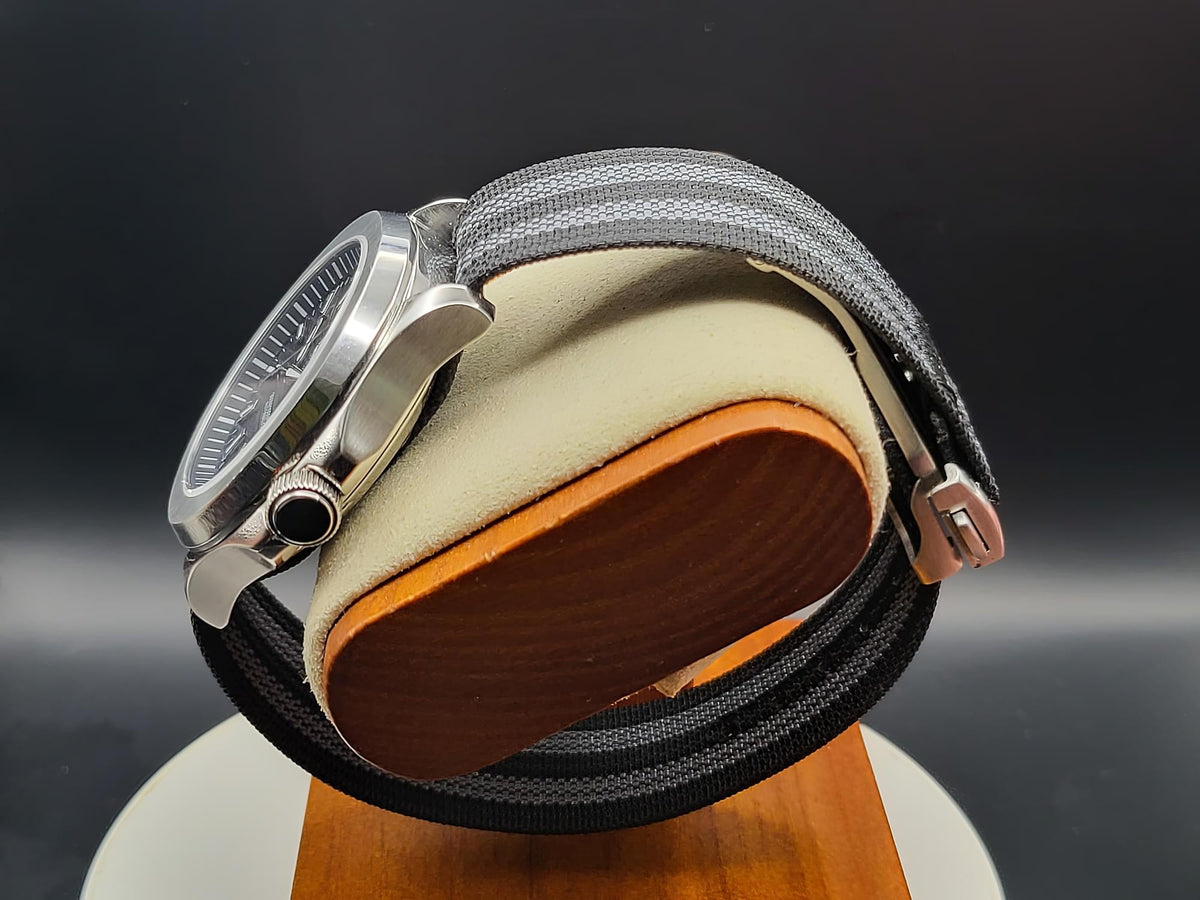 The Alias SJT Original wristwatch, a silver-toned stainless steel watch with a gray and black woven fabric strap, displayed on a beige cushion with a wooden base against a dark background.
