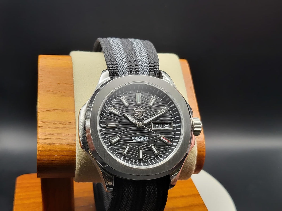 The Alias SJT Original wristwatch with a dark gray wavy dial, silver-toned case, and gray striped fabric strap, displayed on a wooden stand against a dark background.