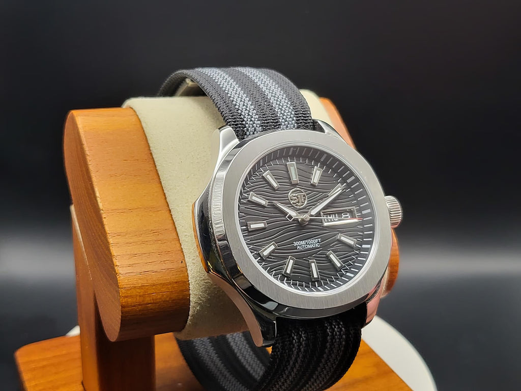 The Alias SJT Original wristwatch with a dark gray dial and a gray-and-black woven strap, displayed on a wooden stand against a dark background.