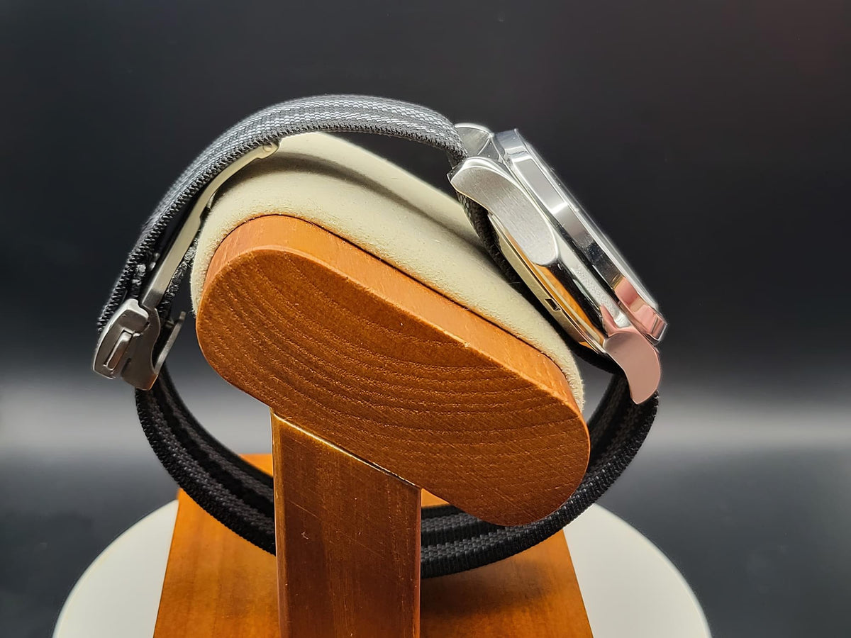 A close-up side view of The Alias SJT Original wristwatch with a silver-toned case, black fabric strap, and light beige leather inlay, displayed on a wooden stand against a dark background.