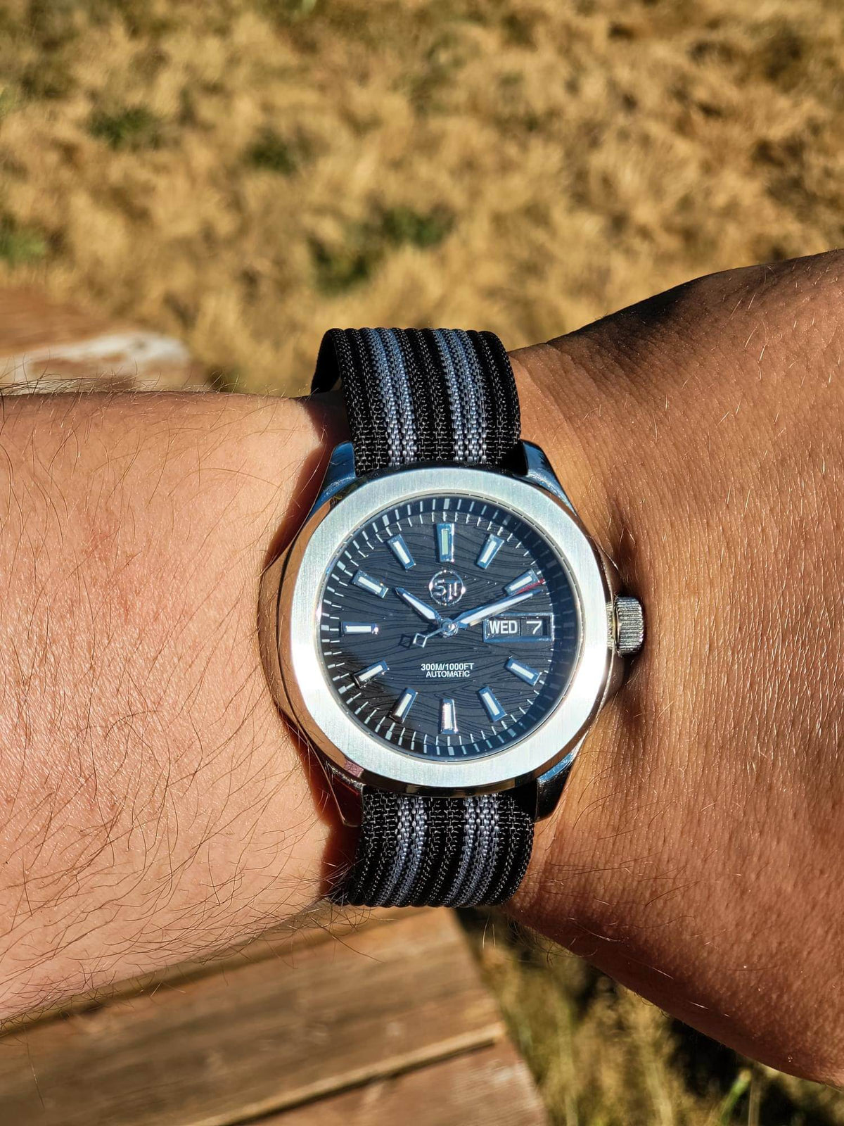 A close-up of a wrist wearing The Alias SJT Original wristwatch watch with a dark blue dial and a black and gray striped nylon strap, set against a blurred outdoor background.