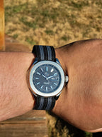 The Alias SJT Original wristwatch with a dark blue dial and a black-and-blue striped fabric strap, worn on a person's wrist against a blurred outdoor background.