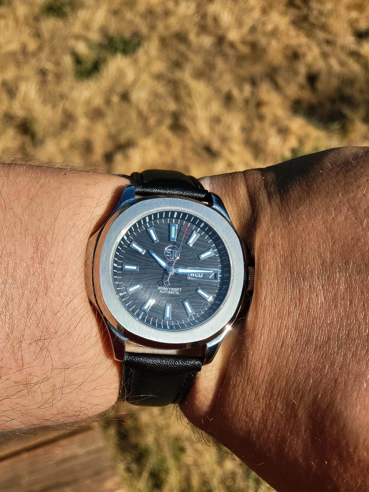 The Alias SJT Original wristwatch with a silver-toned case, blue dial, and black leather strap worn on a person's wrist, photographed outdoors with dry grass in the background.