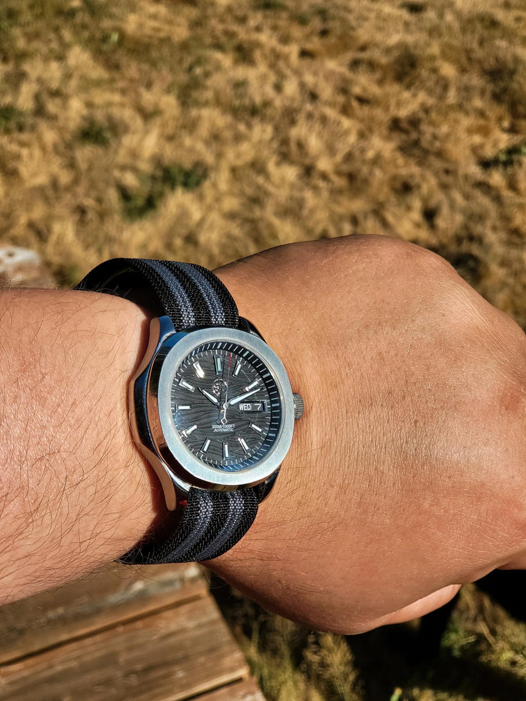 The Alias SJT Original wristwatch with a silver-toned case, black dial, and black-and-gray striped fabric strap, worn on a person's wrist against an outdoor grassy background.