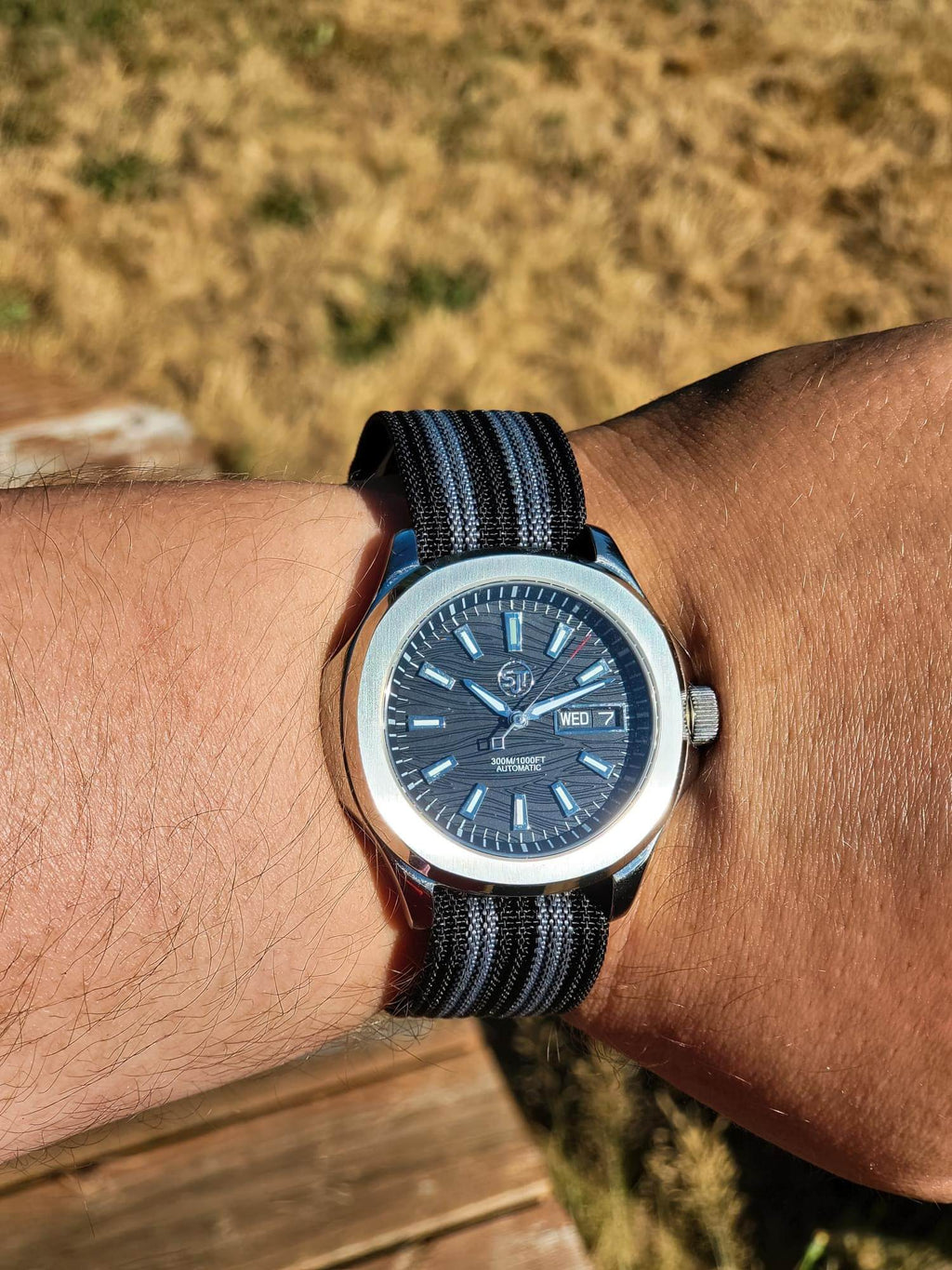 The Alias SJT Original wristwatch with a dark blue dial and a black-and-blue striped fabric strap, worn on a person's wrist against a blurred outdoor background.