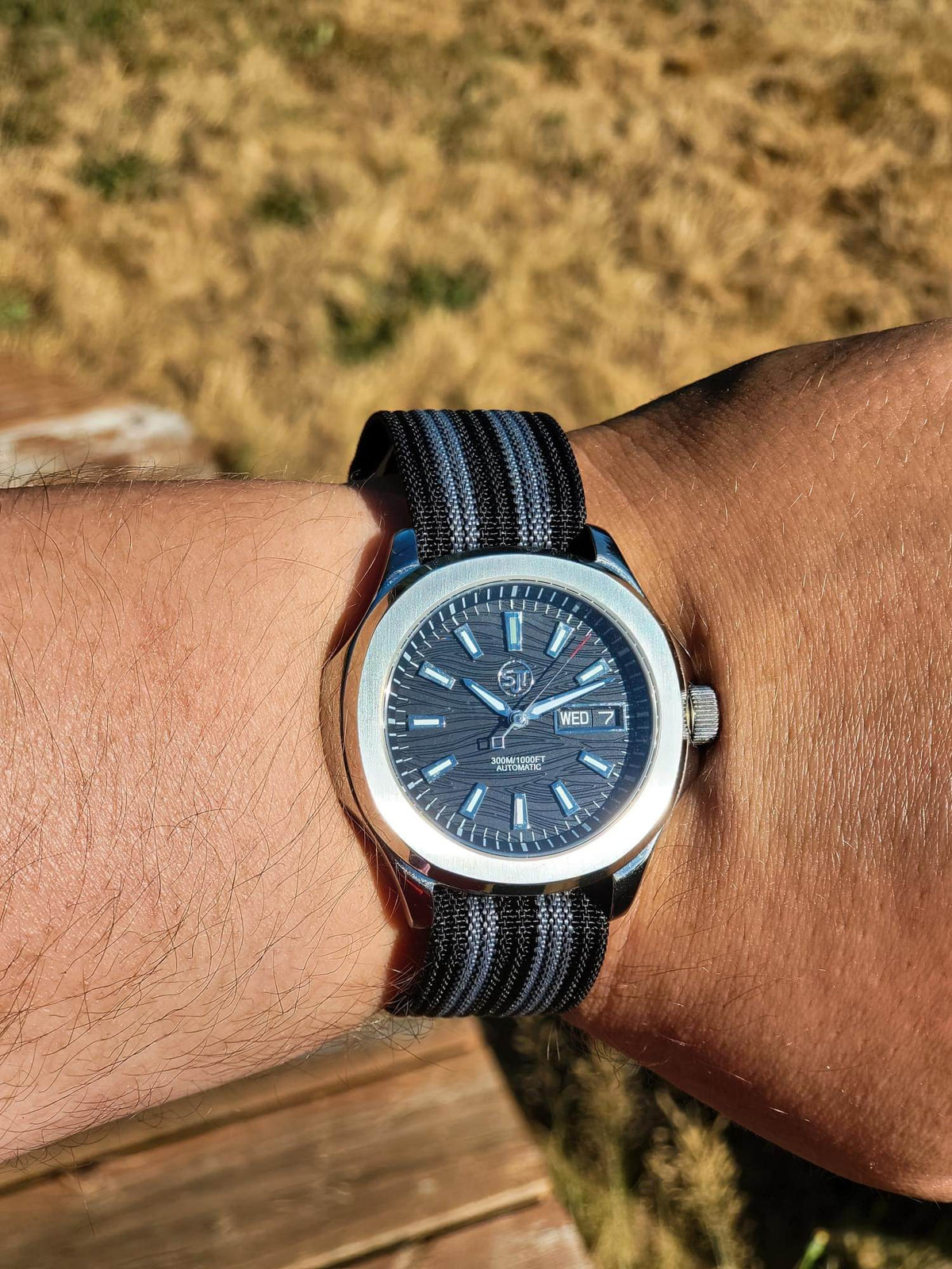 The Alias SJT Original wristwatch with a dark blue dial and a black-and-blue striped fabric strap, worn on a person's wrist against a blurred outdoor background.