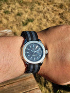 The Alias SJT Original wristwatch with a blue dial and a black-and-blue striped fabric strap, worn on a person's wrist against an outdoor background of dry grass and a wooden deck.