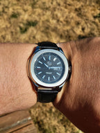 The Alias SJT Original wristwatch with a dark blue dial, silver-toned case, and black leather strap worn on a person's wrist, photographed outdoors with dry grass in the background.