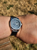 The Alias SJT Original wristwatch with a round silver-tone case, dark gray dial, and black leather strap worn on a person's wrist, photographed outdoors with dry grass in the background.