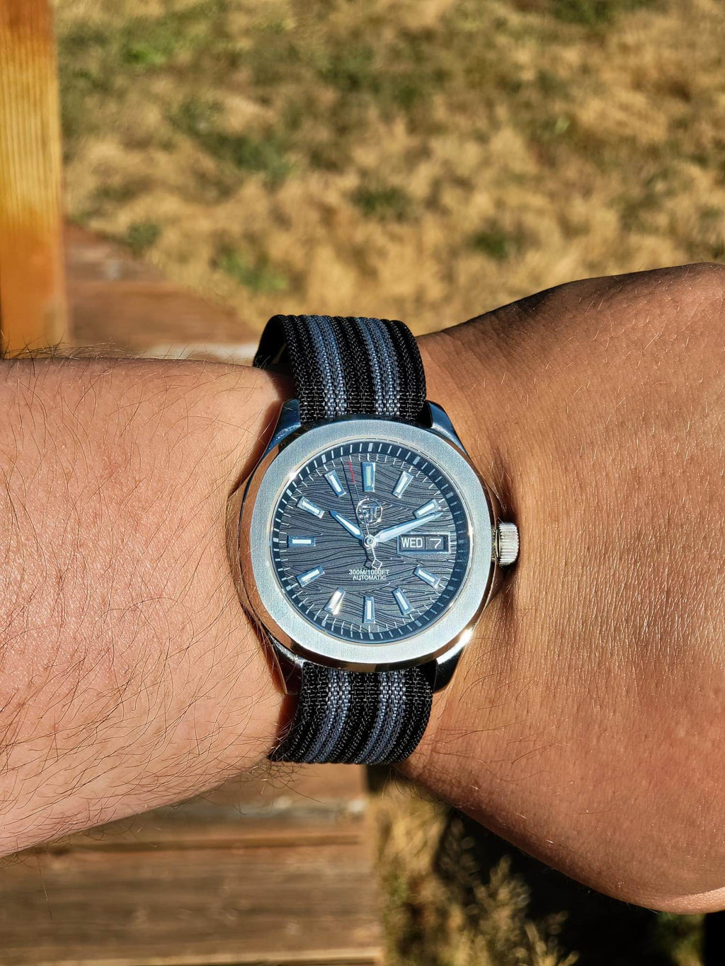 The Alias SJT Original wristwatch with a blue dial and a black-and-blue striped fabric strap, worn on a person's wrist outdoors.