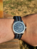 The Alias SJT Original wristwatch with a blue dial and a black-and-blue striped fabric strap, worn on a person's wrist outdoors.
