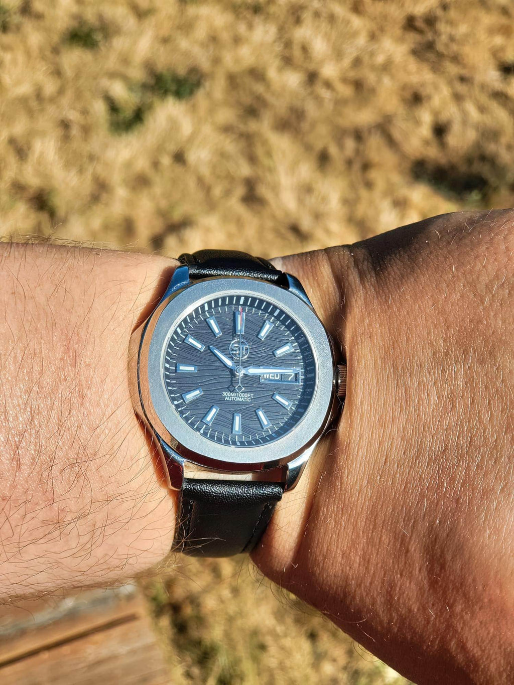 The Alias SJT Original wristwatch with a silver-toned case, blue dial, and black leather strap worn on a person's wrist, photographed outdoors with dry grass in the blurred background.