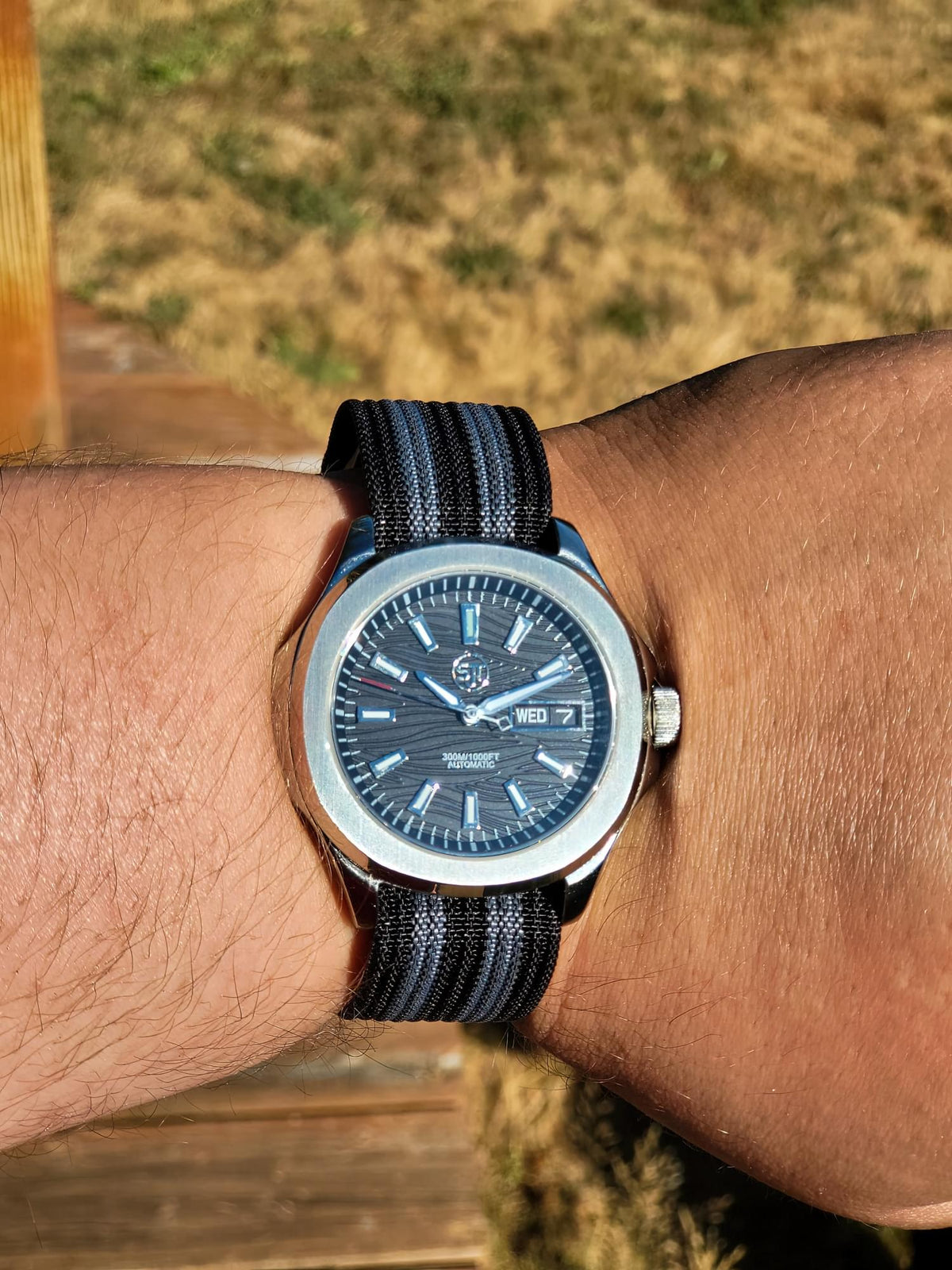 The Alias SJT Original wristwatch with a blue dial and a dark blue-and-black striped fabric strap, worn on a person's wrist outdoors with a blurred wooden deck and grassy background.