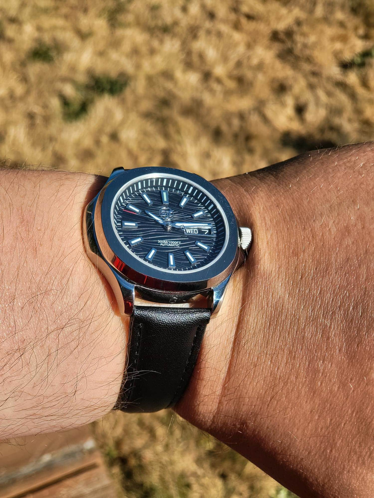 The Alias SJT Original wristwatch with a blue dial, silver-toned case, and black leather strap worn on a person's wrist, photographed outdoors with dry grass in the background.