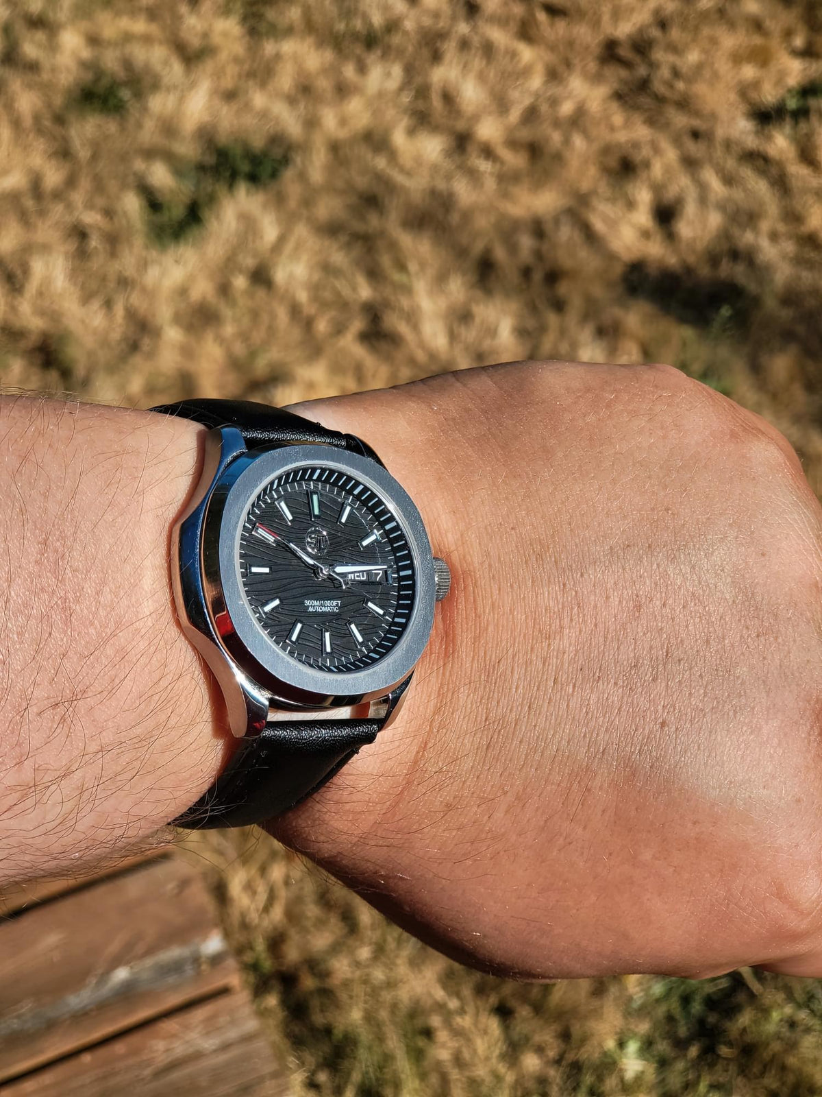 The Alias SJT Original wristwatch with a black dial, silver-toned case, and black leather strap worn on a person's wrist, photographed outdoors against a blurred dry grass background.