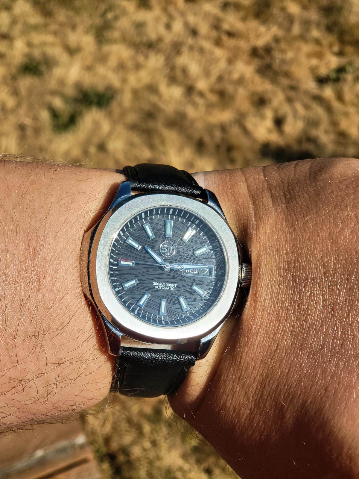 The Alias SJT Original wristwatch with a round silver-tone case, dark blue dial, and black leather strap worn on a person's wrist against a blurred outdoor background.