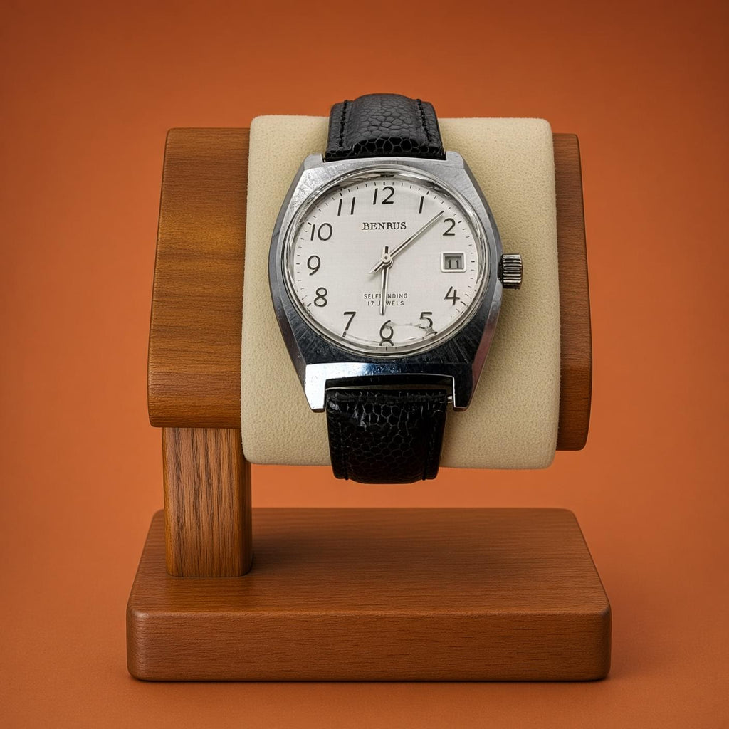 A Benrus wristwatch with a silver-toned case, white dial, and black leather strap, displayed on a beige cushion inside a wooden watch stand against an orange background.