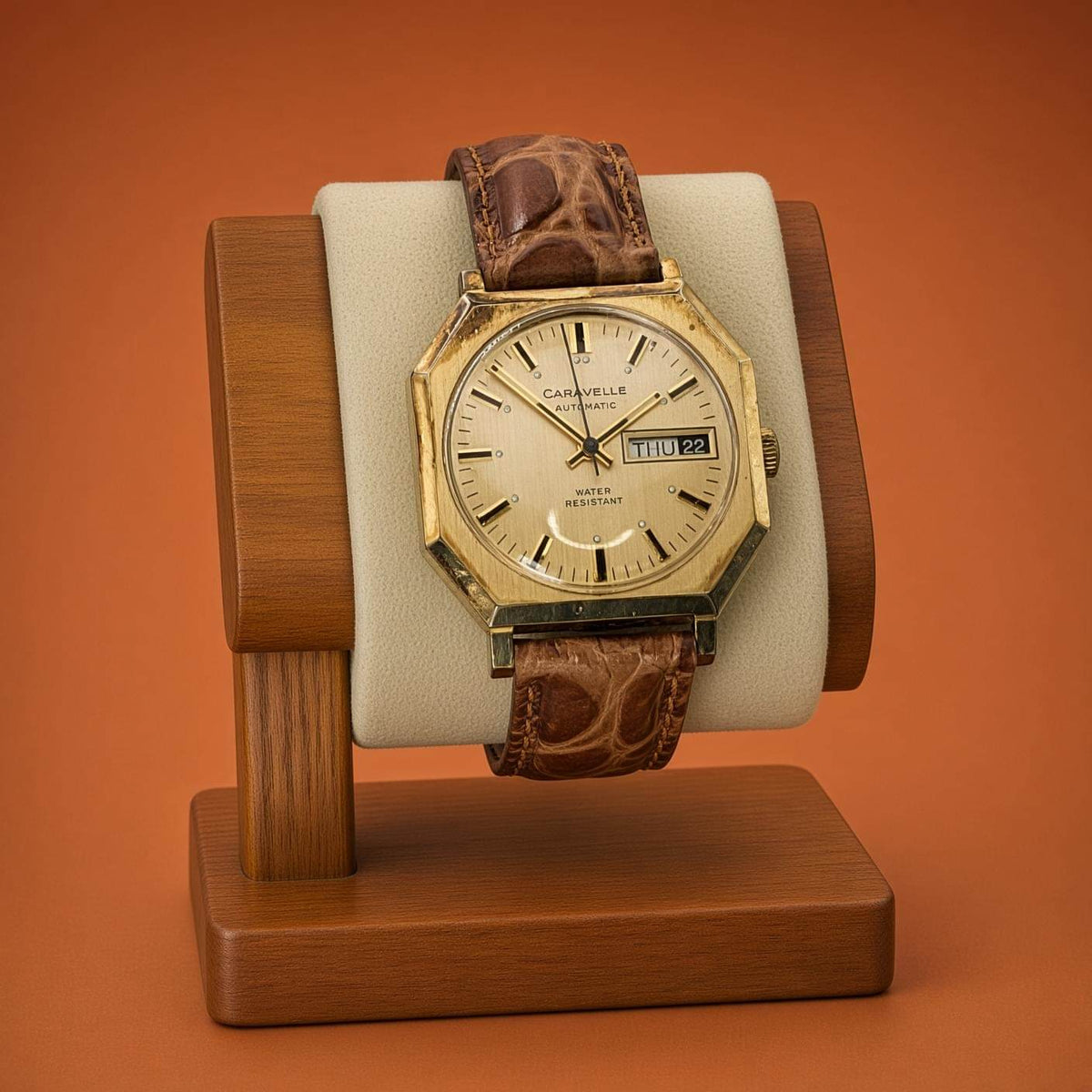 A vintage Caravelle Day-Date watch with a gold-toned octagonal case, cream dial, and brown leather strap, displayed on a wooden stand against a solid orange background.