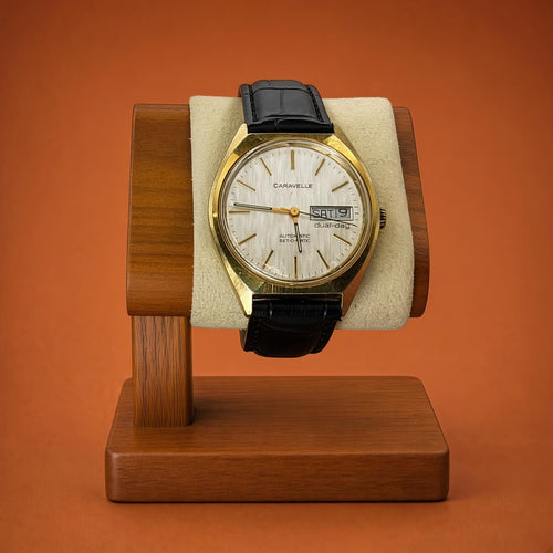 A vintage Caravelle Dual Day watch with a gold-tone case, cream dial, and black leather strap, displayed on a wooden stand against a solid orange background.