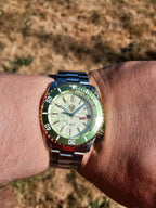 The Coconut SJT Original wristwatch with a green bezel, silver-toned metal bracelet, and a cream-colored dial, shown worn on a person's wrist against a blurred outdoor background.